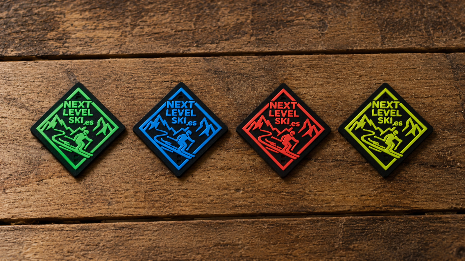 Cuatro parches de goma de Next Level Ski con el logotipo y un esquiador, en verde, azul, rojo y amarillo, colocados sobre una tabla de madera