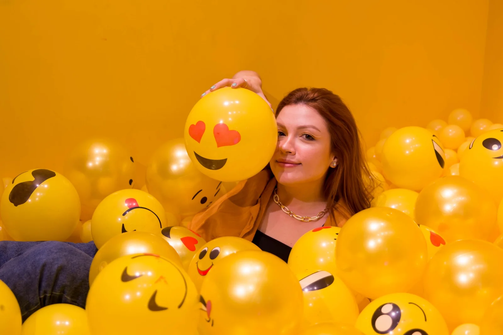 Visitante entre balões amarelos de emoji, segurando um rosto com olhos de coração