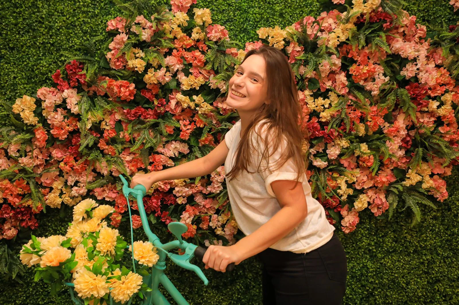 Visitante sorrindo com uma bicicleta turquesa diante de um muro de flores