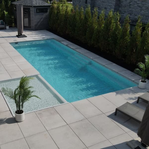 Piscine rectangulaire avec terrasse en béton gris, arbres verts en arrière-plan, abri de piscine, design contemporain