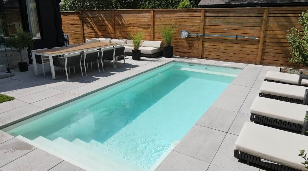 Piscine moderne avec terrasse en béton gris clair, table de salle à manger, chaises noires, clôture en bois et aménagement paysager