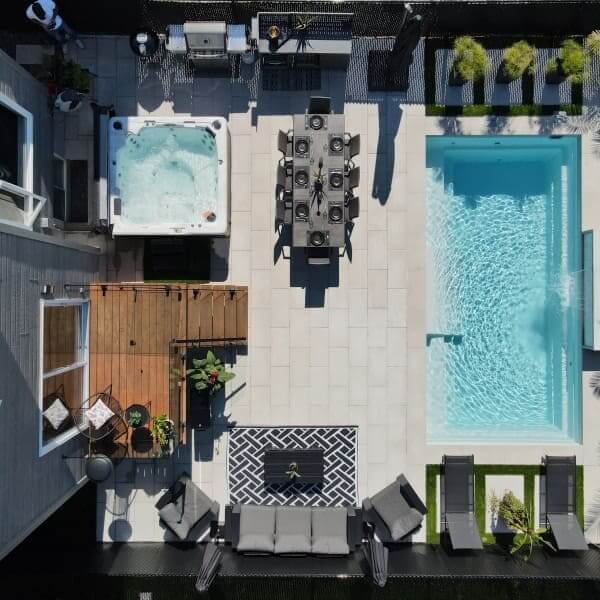 Vue aérienne d'une cour avec piscine creusée, spa, terrasse en pavé blanc, mobilier de détente et aménagement paysager complet