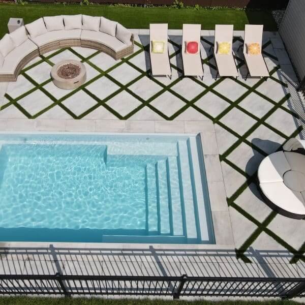 Piscine rectangulaire turquoise avec terrasse en pavé blanc et motif géométrique vert, chaises longues et foyer