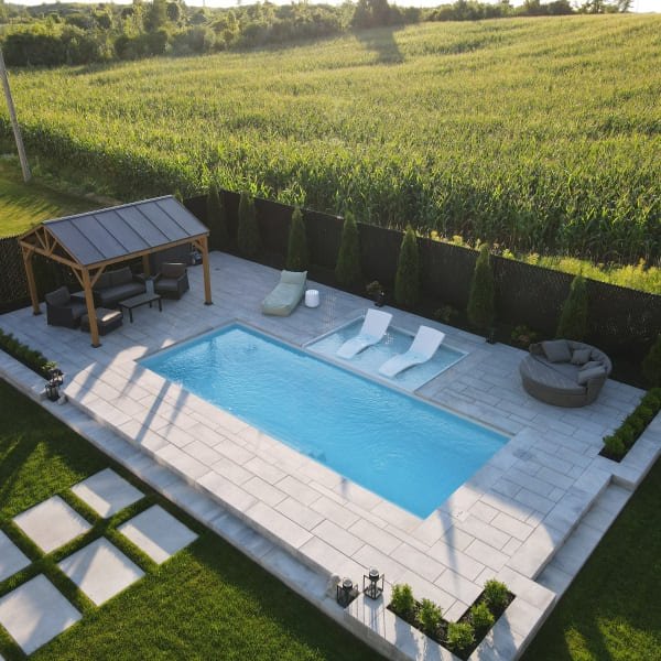 Piscine rectangulaire bleue avec terrasse en pavé blanc, chaises longues, pergola avec panneaux solaires, vue sur champ