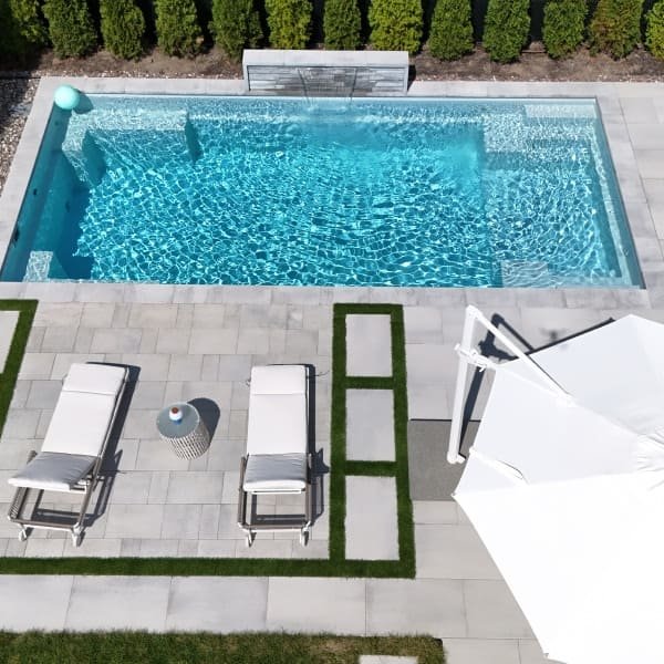 Piscine moderne avec terrasse en béton gris clair, chaises longues blanches, parasol blanc, aménagement minimaliste avec herbe verte