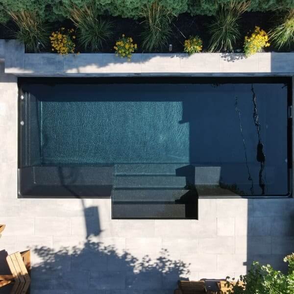 Vue aérienne d'une piscine rectangulaire moderne avec terrasse en béton blanc, clôture noire minimaliste, aménagement paysager avec plantes vertes et fleurs jaunes
