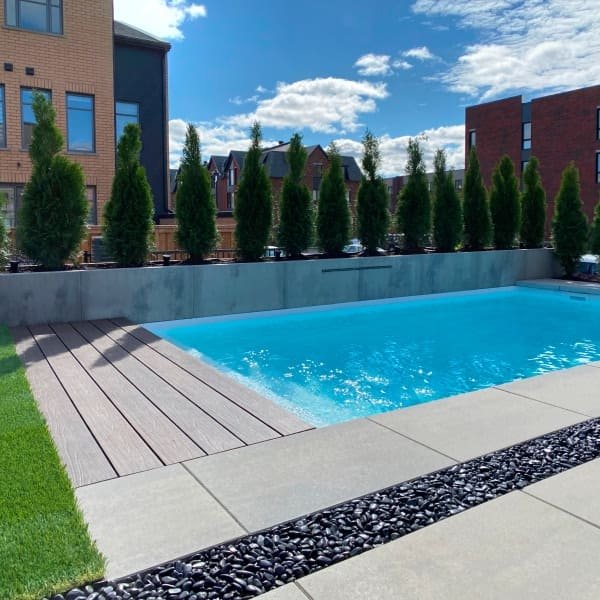Piscine moderne avec terrasse en béton gris, clôture en brique rouge, arbres verts, architecture urbaine