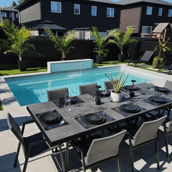 Piscine moderne avec terrasse en pavé gris, table de salle à manger noire avec couverts, chaises longues et palmiers