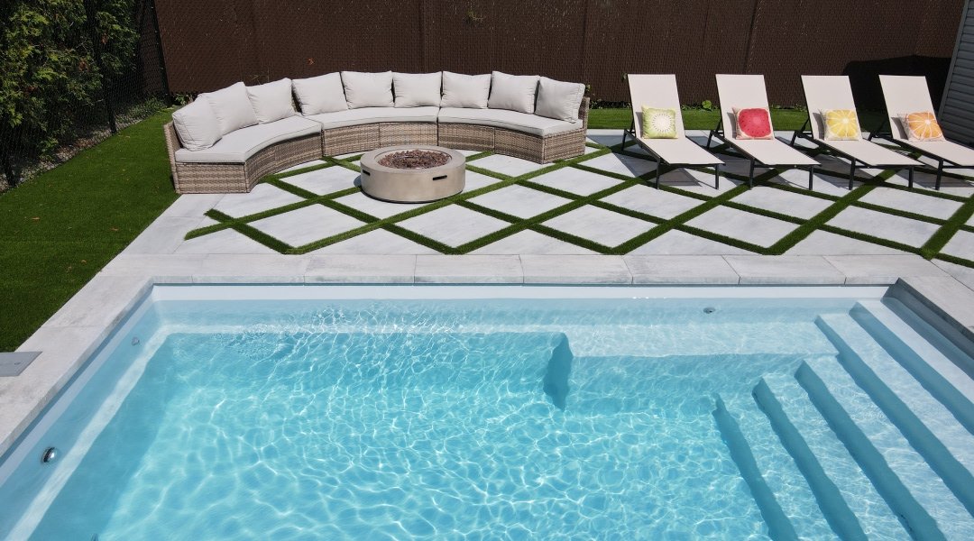 Piscine avec pavé blanc, canapé gris avec coussins, foyer rond et aménagement paysager avec gazon