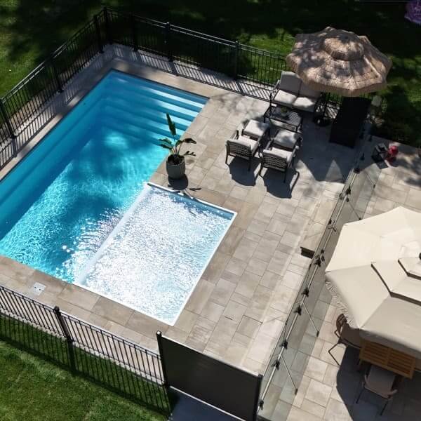 Piscine rectangulaire avec terrasse en béton gris, clôture noire, chaises longues, parasol noir, aménagement paysager avec gazon vert