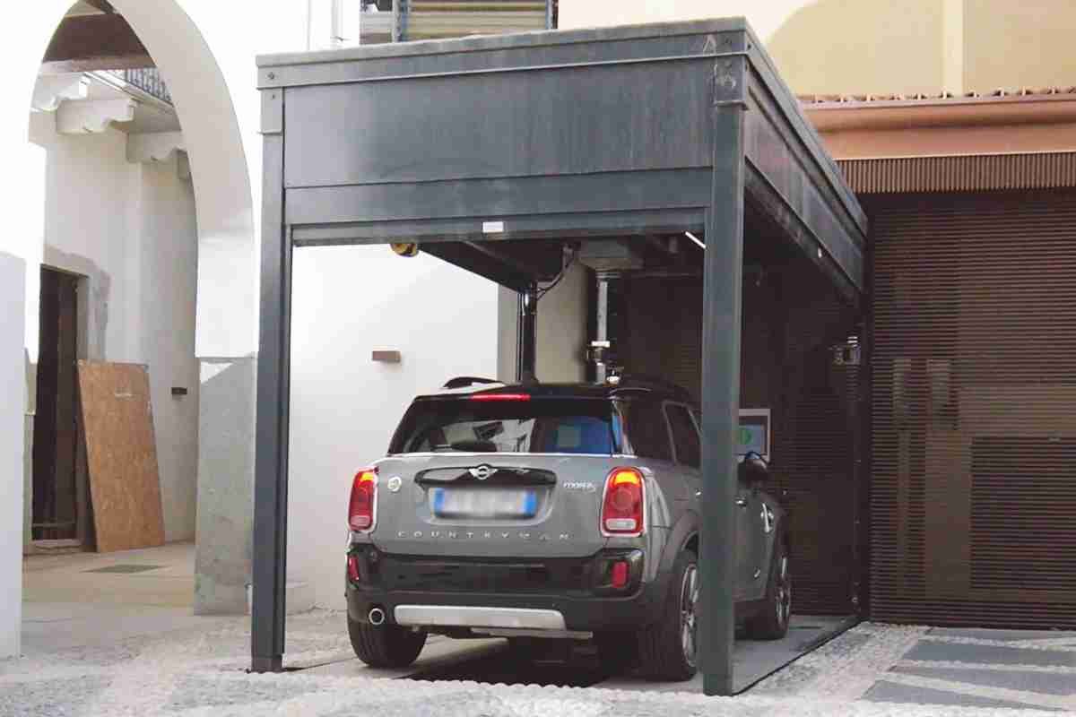 Auto grigia parcheggiata sotto struttura metallica grigia con tetto, cortile interno con edifici residenziali