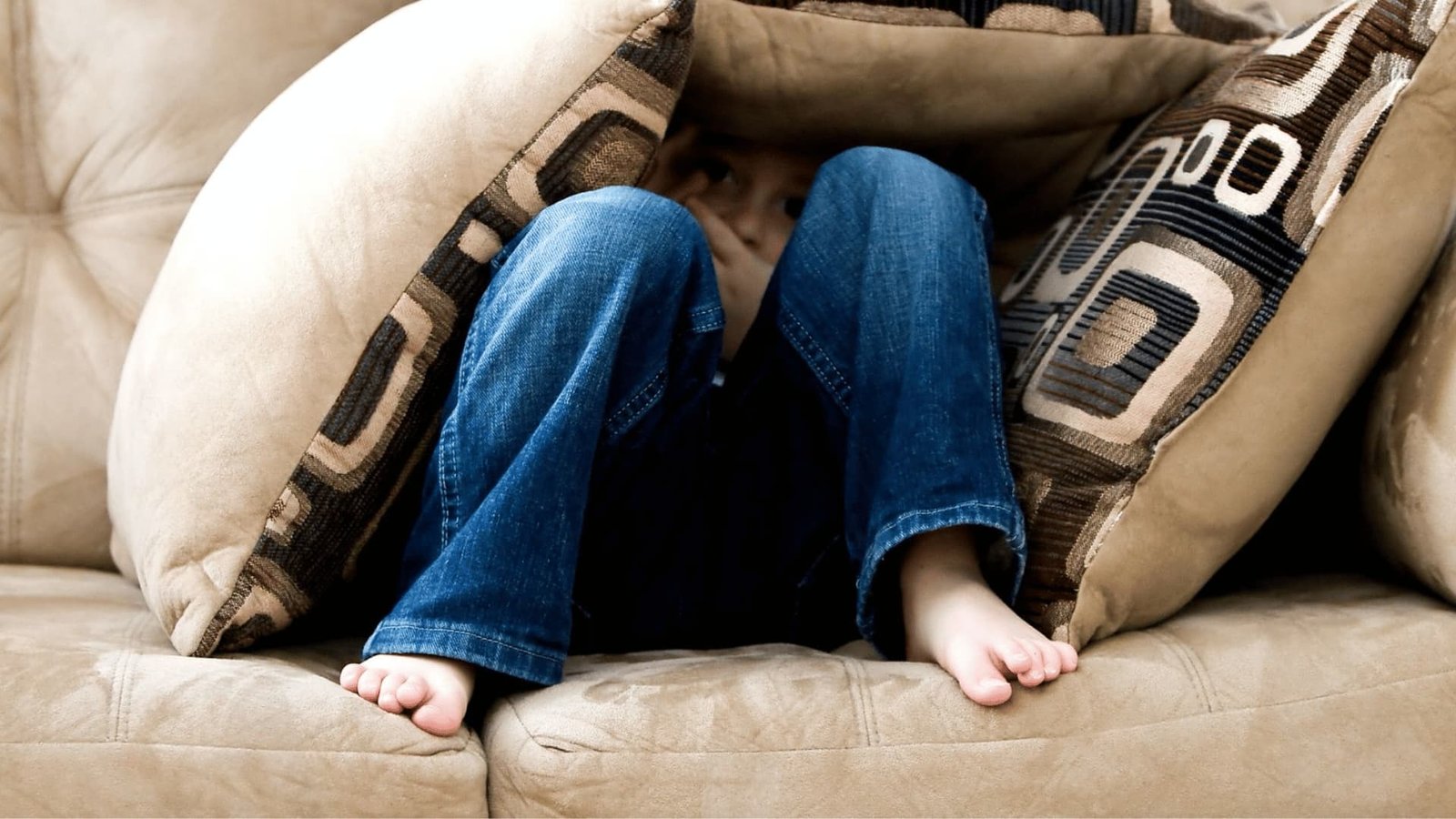 Child hiding under pillows on couch, wearing blue jeans, showing vulnerability