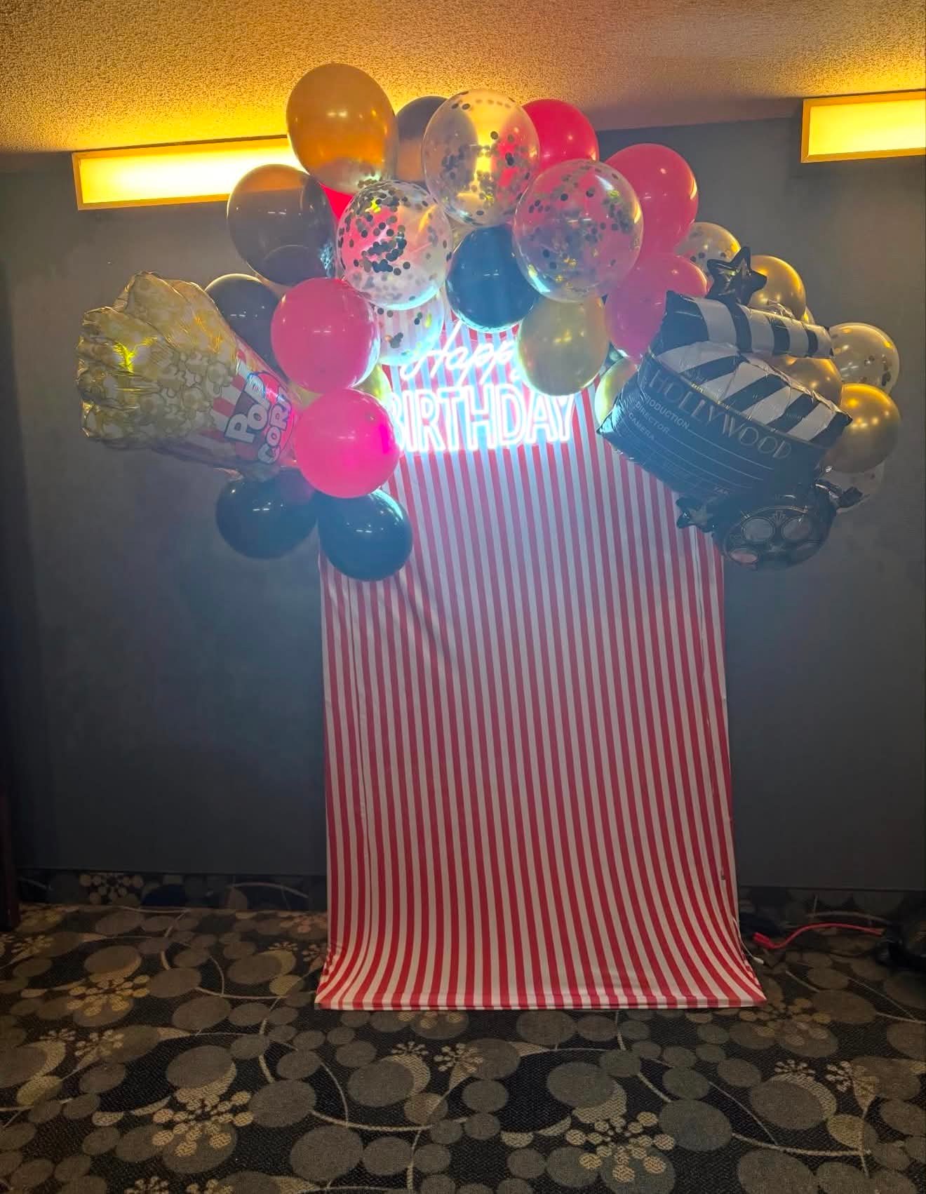 Hollywood-themed balloon arch with popcorn and clapperboard foil balloons over a striped photo backdrop and Happy Birthday neon