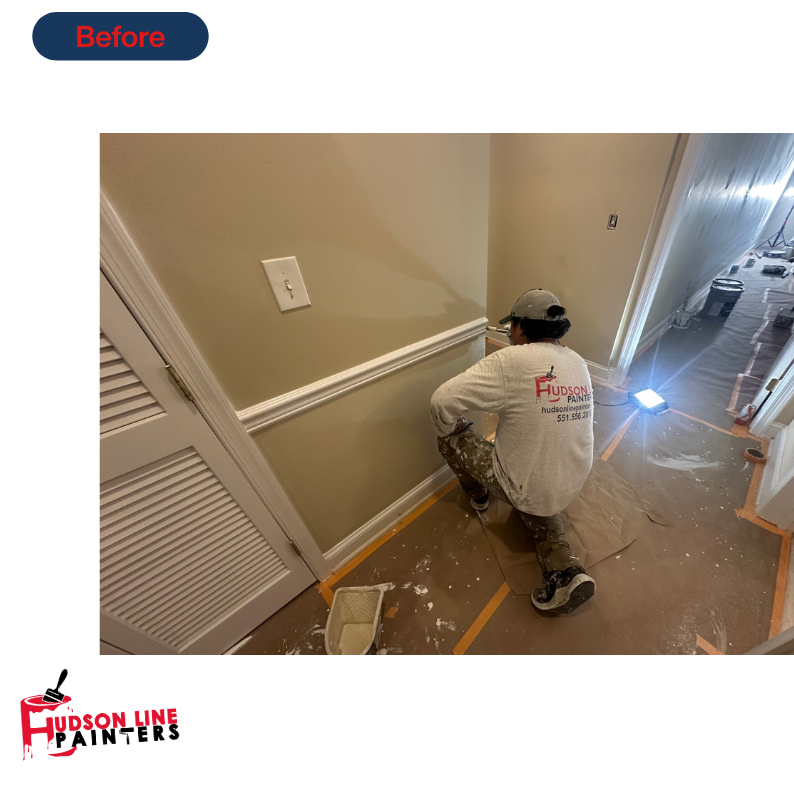 Professional interior painter kneeling to paint a lower wall and chair-rail trim in a residential hallway with floor protection in place