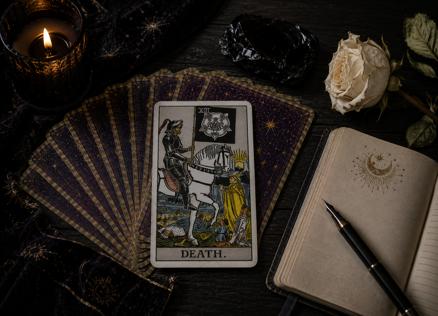 Death tarot card displayed on a dark surface surrounded by a spread of tarot cards, candle, white rose, dried flowers, and an open journal with moon and star illustrations