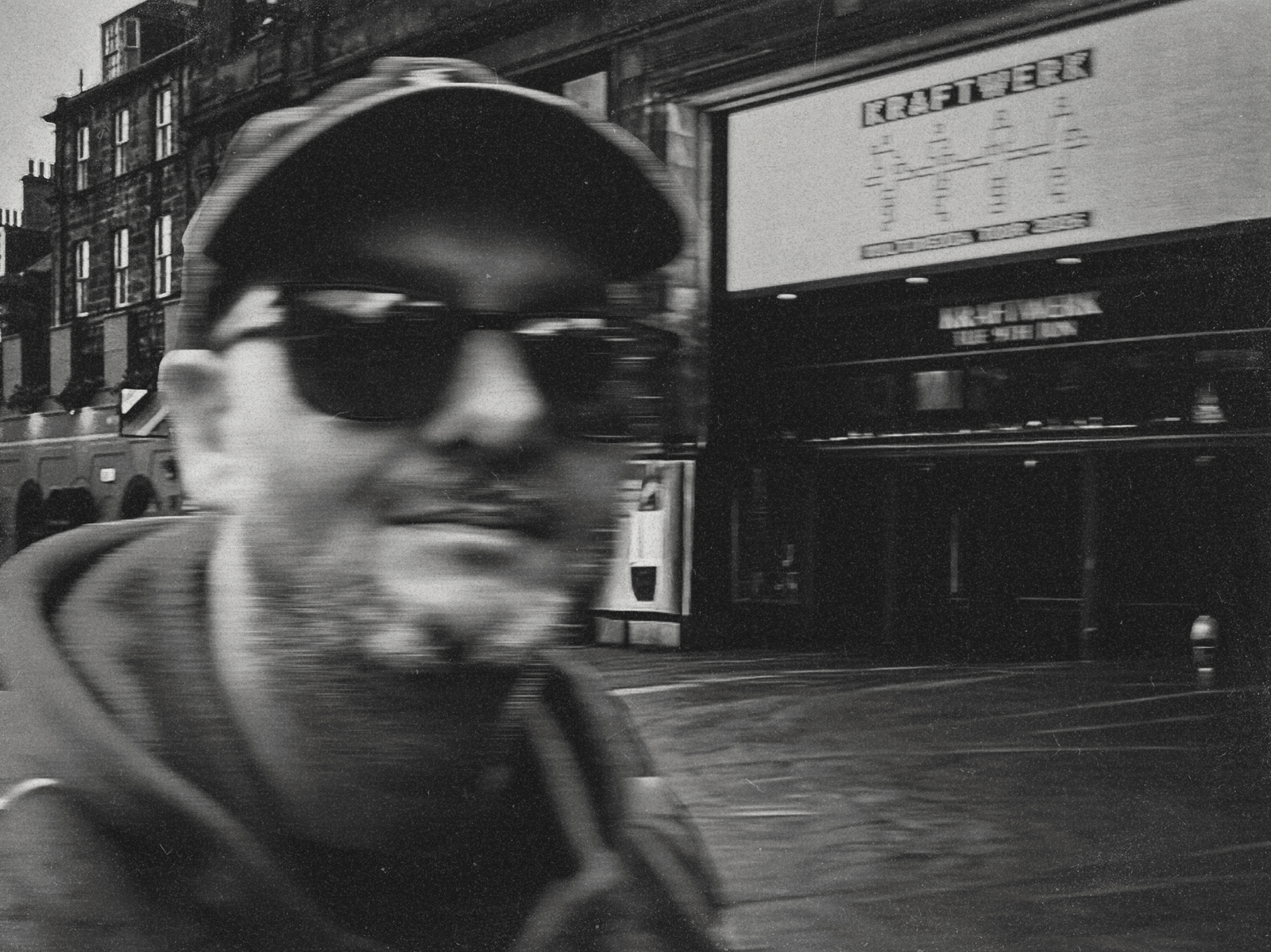 Black-and-white street portrait in front of a Kraftwerk billboard