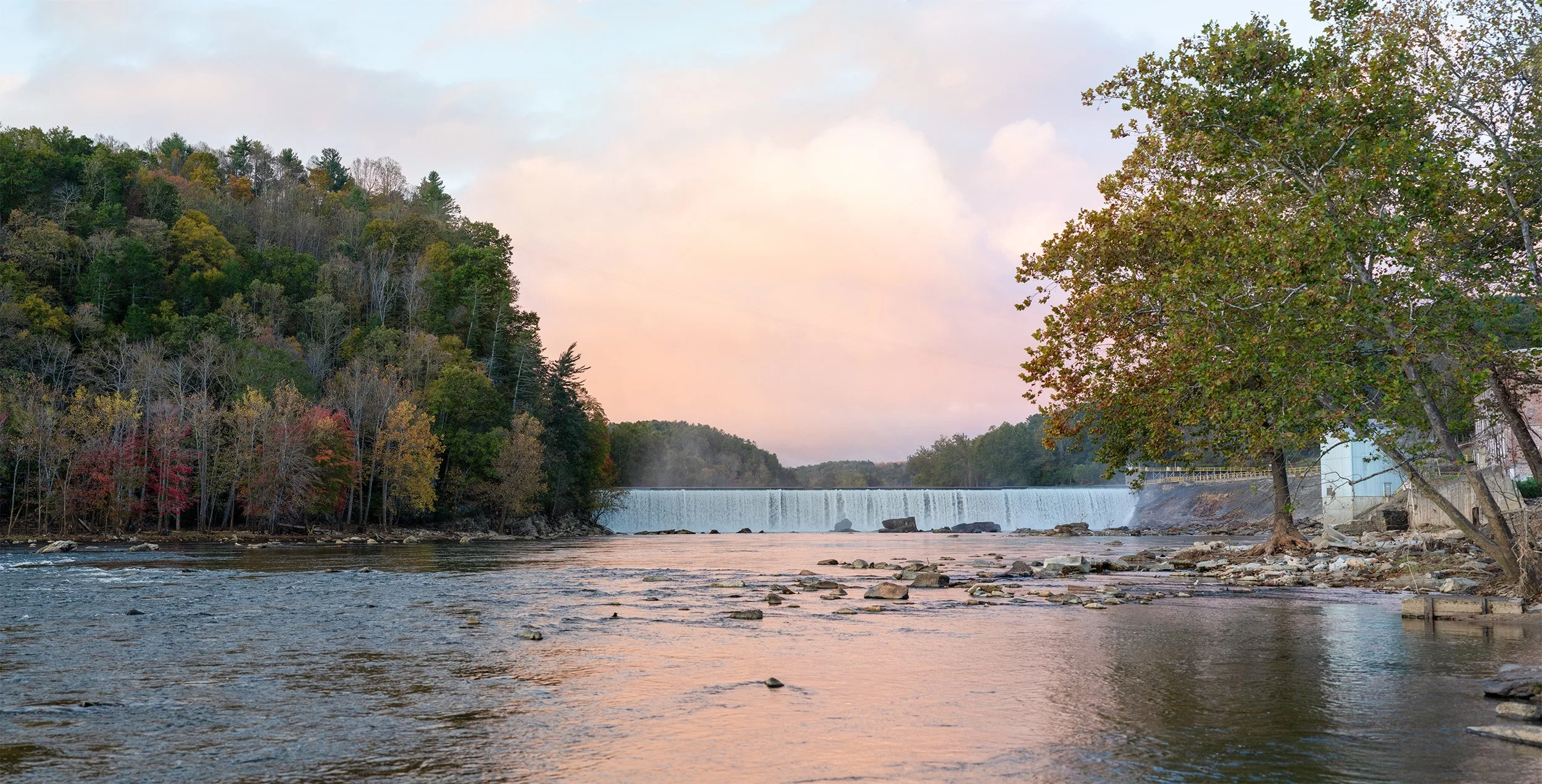 Scenic New River landscape with autumn foliage, river flowing through forested valley with dam/waterfall in background, golden hour lighting