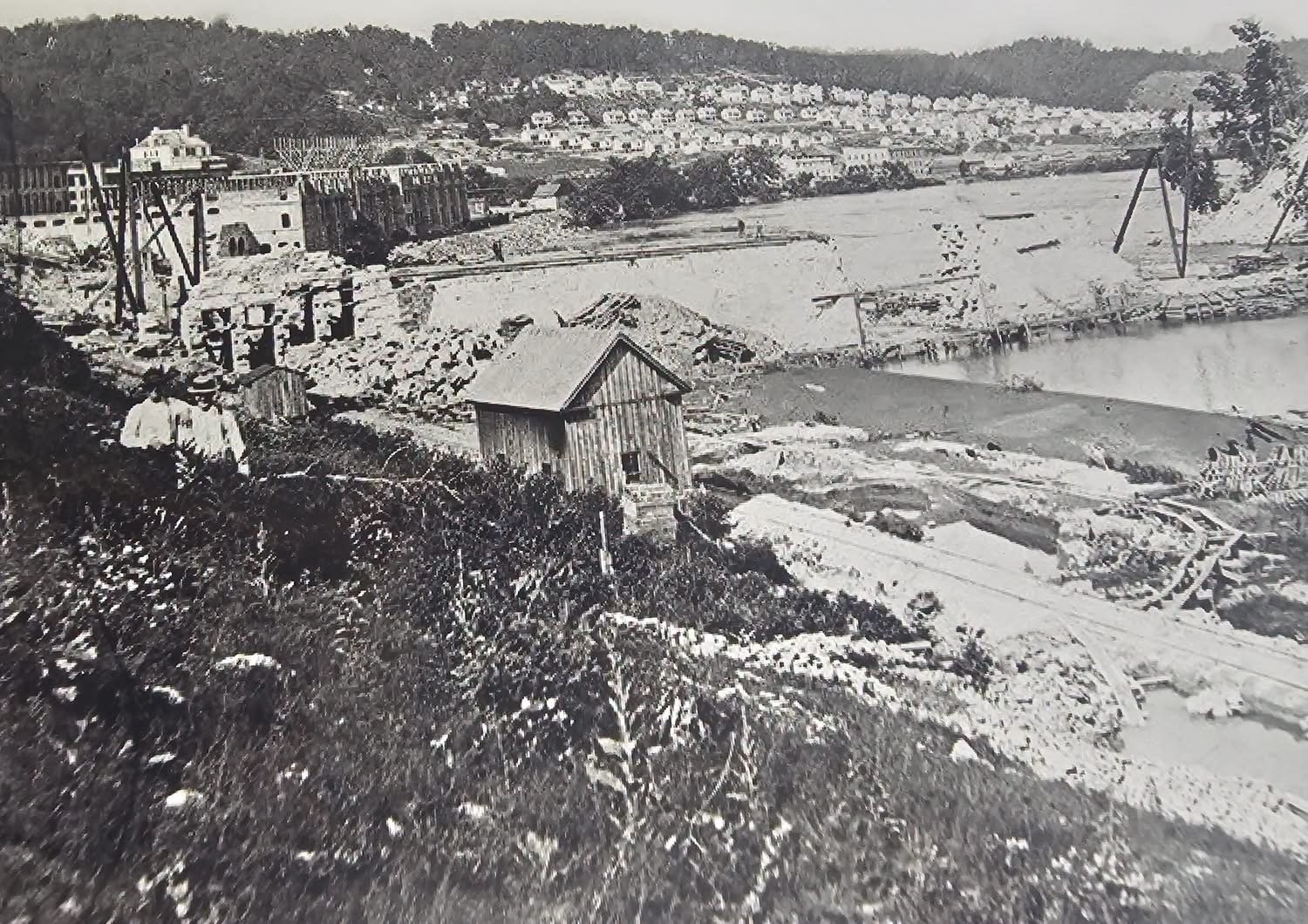Historic black-and-white view of Fries, Virginia, with mill buildings, rubble and construction along the New River, a wooden shed in the foreground, and hillside houses beyond