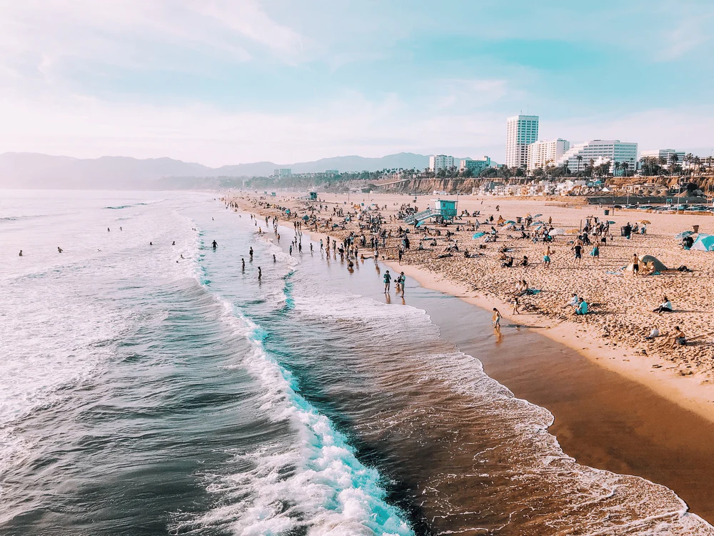 Southern California beach scene with turquoise waves, sandy beach, people swimming and relaxing, coastal buildings and palm trees
