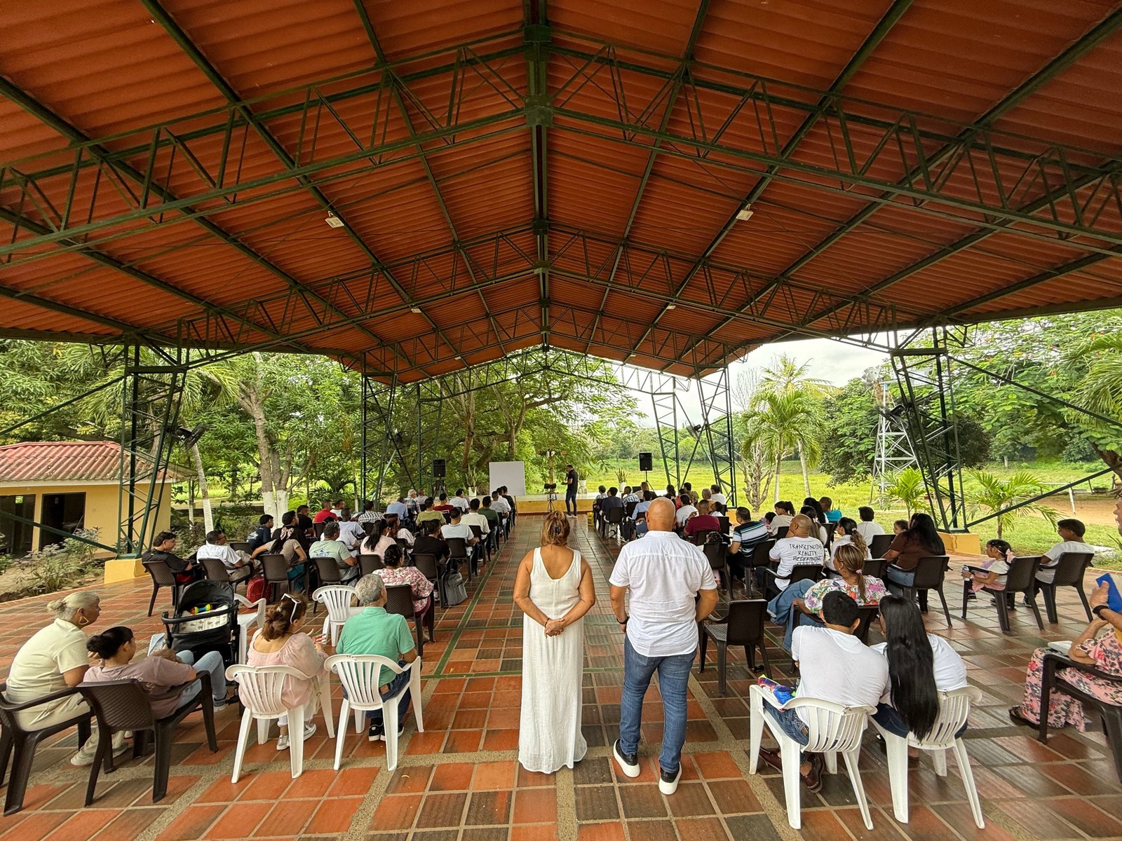 Evento comunitario bajo estructura cubierta con muchas personas sentadas en sillas, árboles verdes de fondo, atmósfera de reunión social y educativa