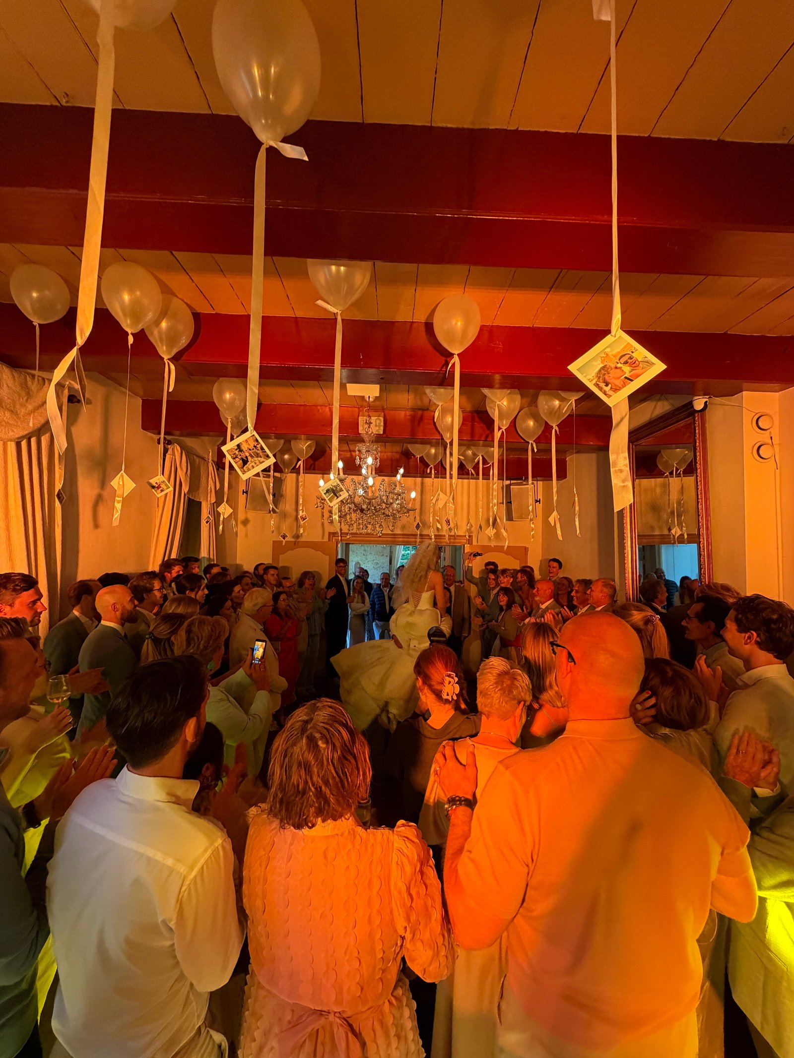 Bruidspaar in het midden van een volle zaal, gasten klappen onder ballonnen en foto’s
