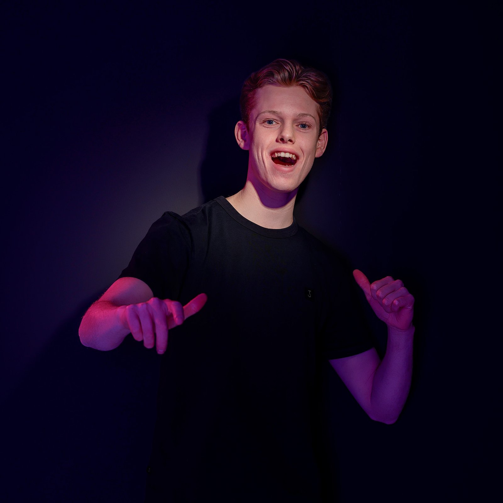 Jongeman in zwart shirt met paarse en roze verlichting op donkere achtergrond, lachend naar de camera
