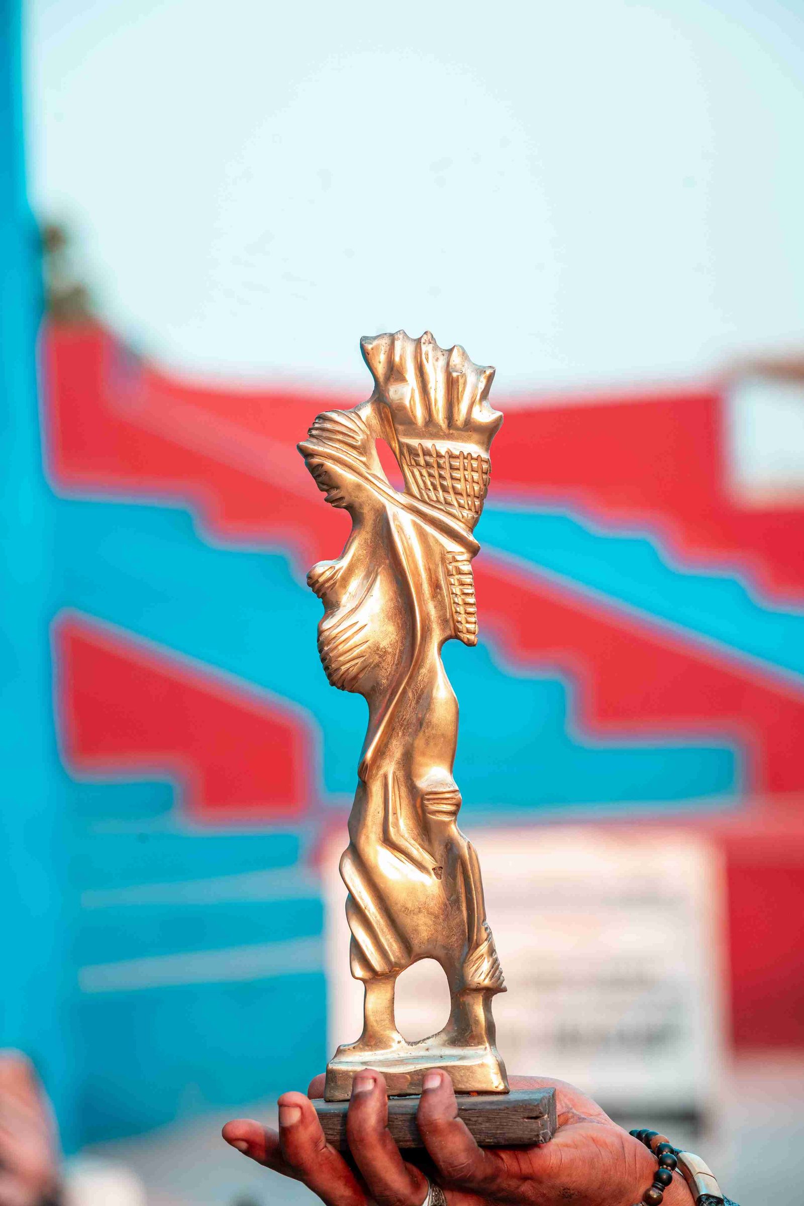 Bronze sculpture of a stylized figure carrying a basket on their head and a child on their back, standing against a blurred background of blue and red geometric shapes.