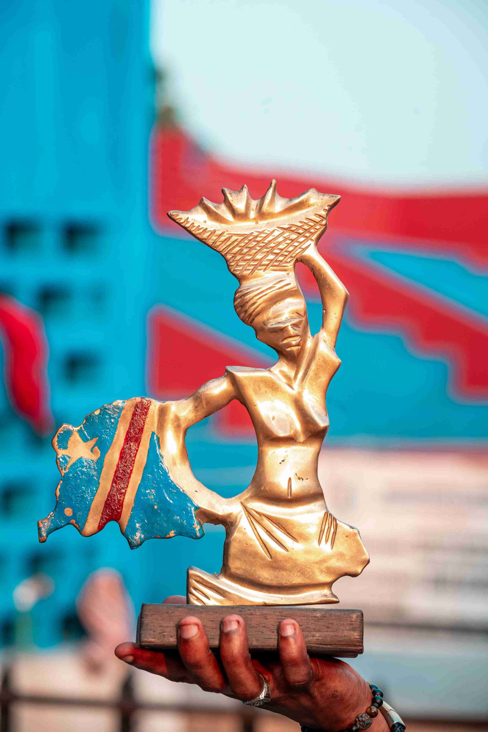 Bronze sculpture of a stylized woman carrying a basket on her head, with a flowing fabric or flag element resembling the flag of Congo attached, held by a hand.