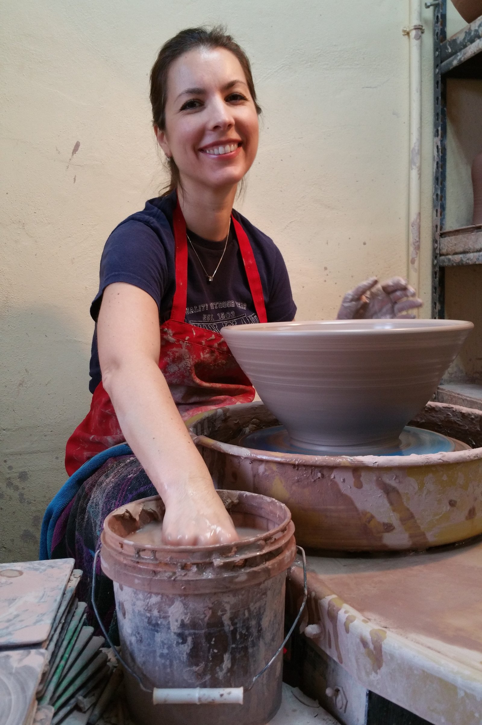 Kimberly Gray smiling in her pottery studio wearing a red apron, surrounded by clay vessels