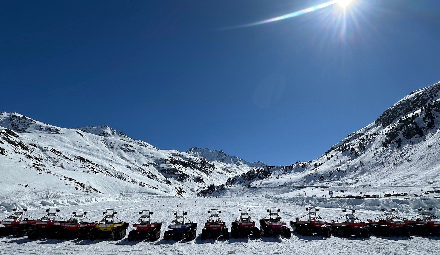 Fourteen Bobsla electric tracked vehicles lined up on snow in Davos, Switzerland, with alpine peaks behind them